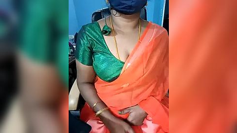 Media: Video of a South Asian woman with medium-dark skin, wearing a green blouse and orange sari, a blue mask, and gold jewelry, seated in a chair.