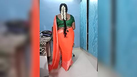 Media: Video of a woman in a green blouse, orange sari, and white garland, walking away in a blue-walled corridor with a patterned door and a bed with a patterned bedspread.