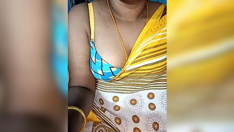 Media: Video of a woman wearing a yellow and blue striped bikini top and a white sari with gold floral patterns. Her skin is medium brown, and she wears gold bangles on her wrist. The background is blurred.