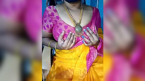 Media: Video of a woman with medium brown skin, wearing a vibrant pink blouse with heart patterns and a yellow sari, adorned with a large golden necklace. Her nails are painted blue. Background is blurred, emphasizing the subject.