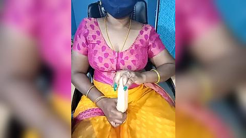 Media: Video of a woman in a bright pink blouse and yellow saree, wearing a mask, holding a white object, seated in a chair against a blue wall.