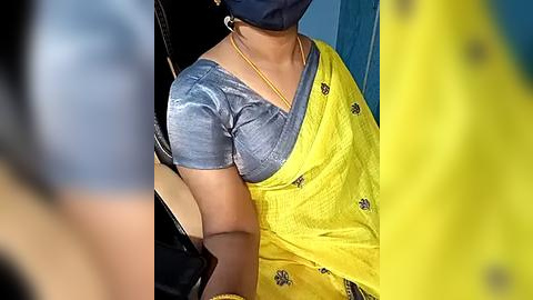 Media: A video shows a woman wearing a yellow saree with floral patterns, a grey blouse, and a blue mask, seated indoors.