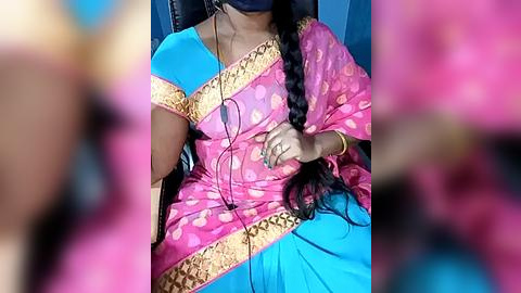 Media: Video of a woman wearing a vibrant pink sari with gold embroidery, blue blouse, and black mask, seated on a black chair, with headphones and a phone on her lap.