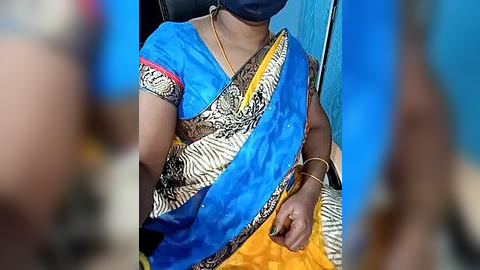 Media: Video of a person wearing a blue sari with a zebra print border, a yellow blouse, and a face mask, seated on a blue chair. The background is blurred.