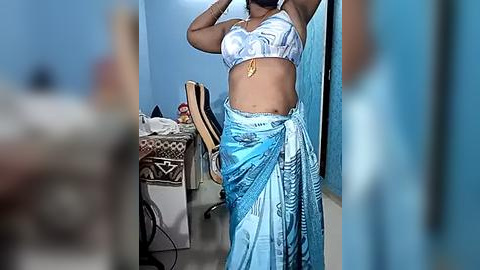 Media: Video of a woman with medium-dark skin, wearing a white lace bra and blue sarong with intricate patterns. She stands in a cluttered, dimly-lit room with a bed, chair, and blue walls.