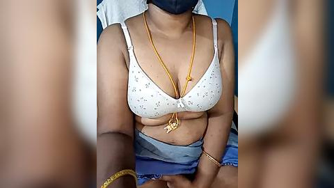 Media: Video of a woman wearing a white bra with small polka dots, a gold necklace, and a blue face mask, sitting in a chair.