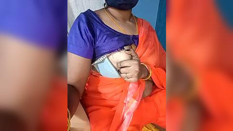 Media: Video of an Indian woman wearing a bright orange saree, blue blouse, and a mask, holding a baby in a white cloth. The background is a plain blue wall.