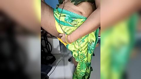 Media: Video of a person in a green and yellow patterned skirt, bending over to tie a yellow shoelace, in a modern office with a desk and chair visible in the background.