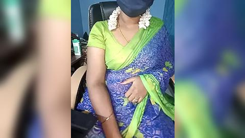 Media: Video of a woman in a blue sari with a green border, seated in an office chair, wearing a blue headscarf and a black mask.