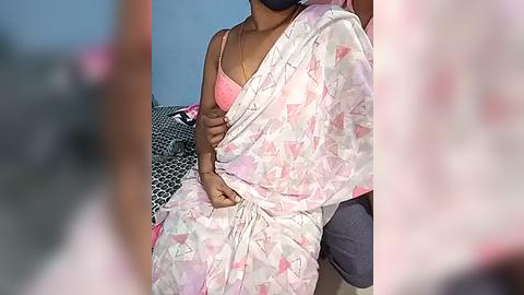 Media: Video of a South Asian woman in a pink sari with geometric patterns, wearing a matching pink bra, sitting on a bed with a black and white patterned pillow.