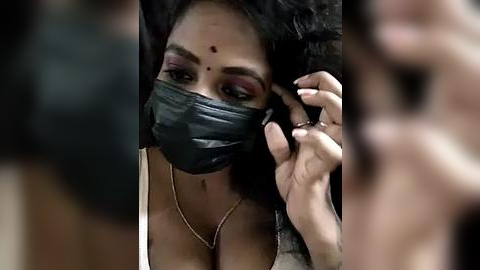 Media: Video of a woman with medium-dark skin, wearing a black face mask, adjusting her makeup with her right hand. She has long, wavy black hair and is wearing a white top. The background is blurred.