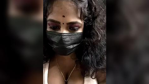 Media: Video of a woman with medium brown skin, long curly black hair, and a black face mask, wearing a white top, and a gold necklace, with a prominent red bindi on her forehead.