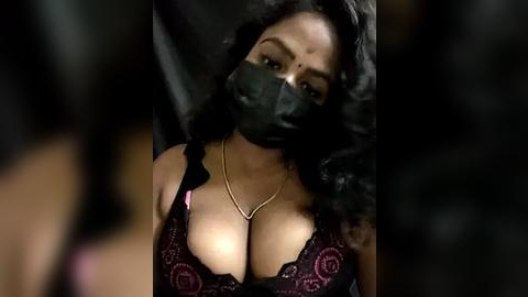 Media: Video of a woman with medium brown skin, wearing a black lace bra with pink floral embroidery, and a black face mask, lying on a black sheet.