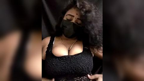 Media: A video of a dark-skinned woman with curly hair, wearing a black lace top that reveals cleavage, a black mask, and a gold necklace, against a blurred background.