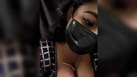 Media: Video of a woman with dark skin, wearing a black face mask, black patterned blouse, and gold necklace. Her eyes are partially visible, focused on a blurred background.