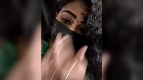 Media: Video of a South Asian woman with dark skin, covered in a black mask, wearing a green sari, with black hair, focused expression, blurred background.