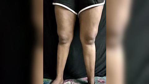 Media: Video of a woman's legs in black shorts with white trim, standing on a floral-patterned rug against a dark background.