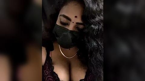 Media: A close-up video of a woman with medium brown skin, wearing a black face mask, revealing her prominent, full lips and large breasts, partially visible through a lacy black top. Her long, wavy black hair cascades around her shoulders.