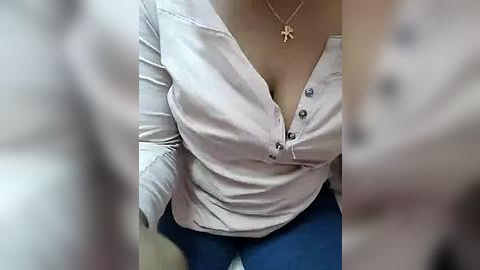 Media: Video of a woman with light skin wearing a white, button-up blouse that's unbuttoned, revealing cleavage. She has a gold necklace and blue jeans. Background is blurred.