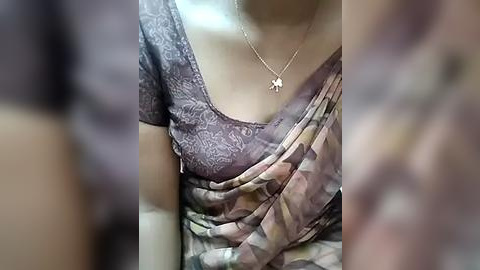 Media: Video of a woman's upper body wearing a floral-patterned blouse and a draped, multicolored sari, with a gold pendant necklace. Blurred background.