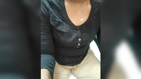 Media: Video of a woman with light brown skin wearing a black button-up blouse, beige pants, and a gold necklace with a cross pendant. The background is blurred and dark.