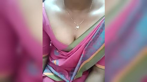 Media: Video of a woman in a bright pink and green sari with a low neckline, revealing cleavage. She wears a gold necklace with a small pendant. Background blurred, focusing on her attire.