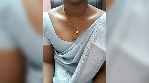 Media: Video of a South Asian woman with medium brown skin, wearing a light blue saree with intricate embroidery, a gold necklace with a cross pendant, and seated on a black leather chair.