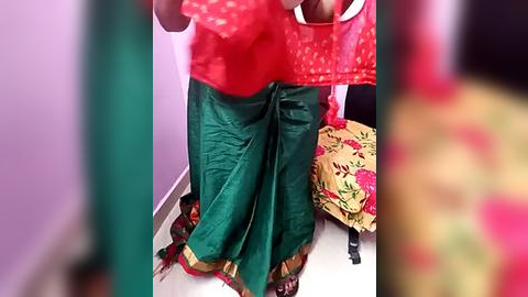 Media: Video of a person in a bright red, polka-dotted blouse and green, floral-patterned skirt, with a purple wall and a yellow, flower-patterned cushion in the background.