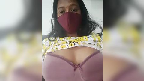 Media: Video of a woman with long black hair, wearing a red face mask, a yellow and white patterned shirt lifted to reveal a purple lace bra, and a maroon top. Background is blurry with indistinct objects.