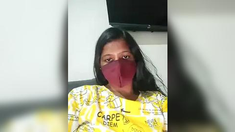 Media: Video of a woman with long black hair, wearing a red face mask and a yellow shirt with \"Carpe Diem\" print, seated indoors with a black TV mounted on a white wall in the background.