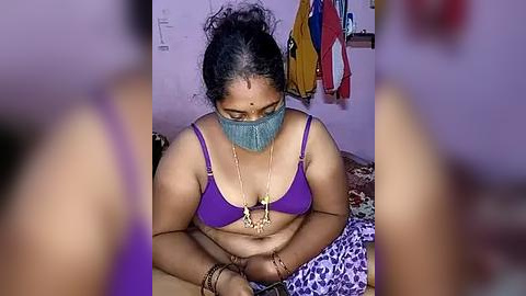 Media: A video of an overweight woman with medium brown skin wearing a purple bra, gray mask, and multiple necklaces, sitting cross-legged on a leopard-print bed.