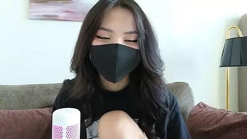 Media: Video of an Asian woman with long black hair, wearing a black face mask and black shirt, sitting on a brown sofa with a white speaker and a black lamp in the background.