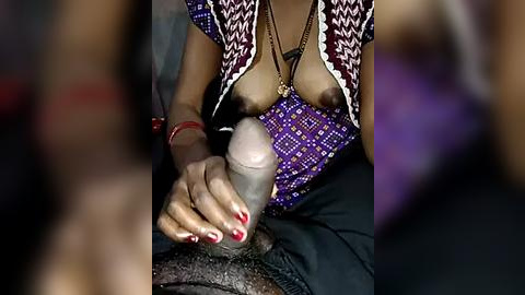 Media: A video of a woman with medium brown skin, wearing a purple patterned top, revealing her large breasts. She has red-painted nails and is using a pink dildo on a man's erect penis.