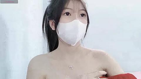 Media: Video of an Asian woman with long black hair, wearing a white mask, red gloves, and a red top, standing in a white room, with visible skin marks and a small butterfly tattoo on her chest.