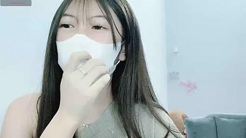 Media: A video of an East Asian woman with long black hair, wearing a white surgical mask, a gray top, and a ring, in a light-colored room with a white wall and pink decor.