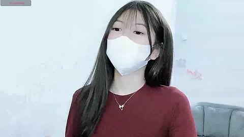 Media: Video of an East Asian woman with long black hair, wearing a white medical mask and a maroon sweater, standing indoors in a soft-focus, light-colored room.