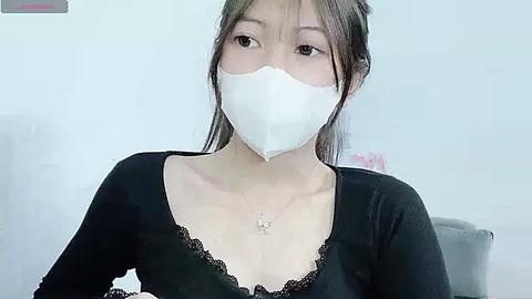 Media: A video of an East Asian woman with light skin, brown hair in a ponytail, wearing a white surgical mask, black lace-trimmed top, and a delicate necklace.