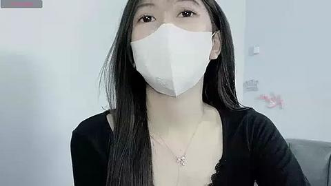 Media: Video of an Asian woman with long black hair, wearing a white face mask and a black lace-trimmed top. She has fair skin and is indoors with a white wall and a gray couch in the background.