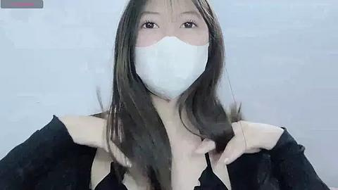 Media: Video of an East Asian woman with long dark hair, wearing a white face mask and a black top, against a plain white background.