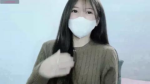 Media: Video of an Asian woman with long black hair, wearing a white mask, green sweater, and sitting indoors. She appears to be adjusting her mask.