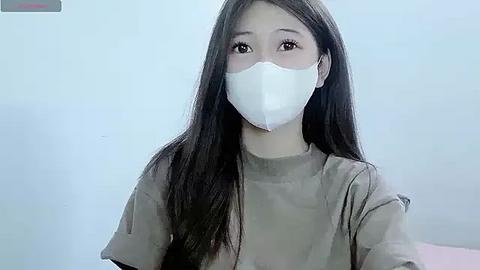 Media: Video of an Asian woman with long black hair wearing a white face mask, beige top, and pale blue background.