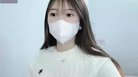 Media: Video of an East Asian woman with long dark hair, wearing a white face mask and a white sleeveless top with a small black logo. Background is a plain, light-colored wall.