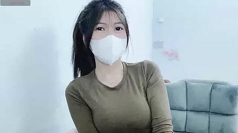 Media: Video of an Asian woman with long black hair in a ponytail, wearing a beige long-sleeved shirt and a white surgical mask, sitting indoors on a gray couch.