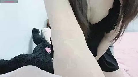 Media: Video of a young woman with fair skin, long brown hair, wearing a black dress, lying on a bed with a fluffy black teddy bear beside her.