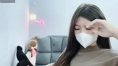 Media: Video of a young Asian woman with long, straight black hair, wearing a white mask, sitting on a gray sofa, touching her face, surrounded by stuffed animals.