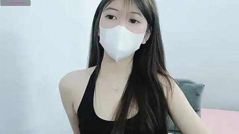 Media: Video of an East Asian woman with long black hair, wearing a white mask, black tank top, and necklace, in a white-walled room.