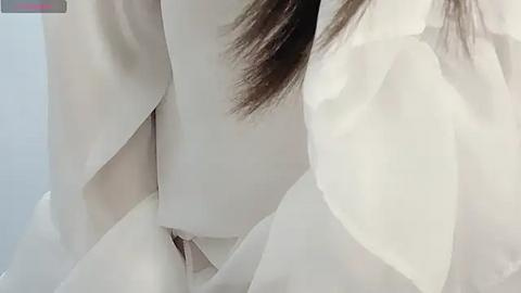 Media: A close-up video of a person's face, partially obscured by white fabric, possibly a surgical mask or veil, with long brown hair visible on the right side. The background is a soft, neutral color.