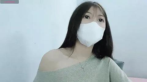 Media: Video of an Asian woman with long black hair, wearing a white mask and a light grey off-shoulder sweater, indoors against a plain white wall.