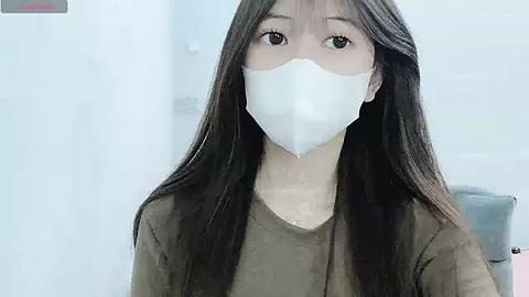 Media: A video of a young Asian woman with long black hair, wearing a white surgical mask, olive green shirt, and seated indoors.