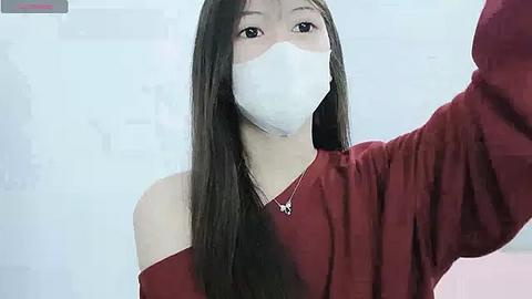 Media: Video of an East Asian woman with long black hair, wearing a white face mask, red off-shoulder sweater, and a delicate necklace, taking a selfie in a softly lit room.